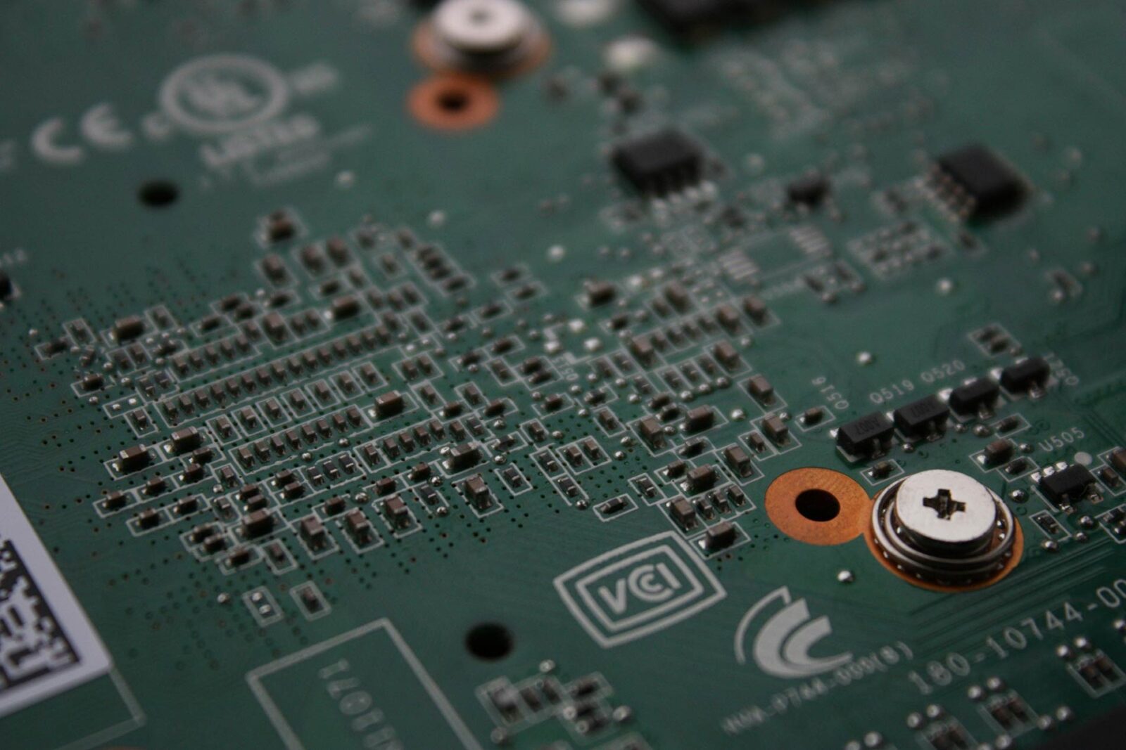 A close-up of a green circuit board with various electronic components, solder points, and a metallic screw.